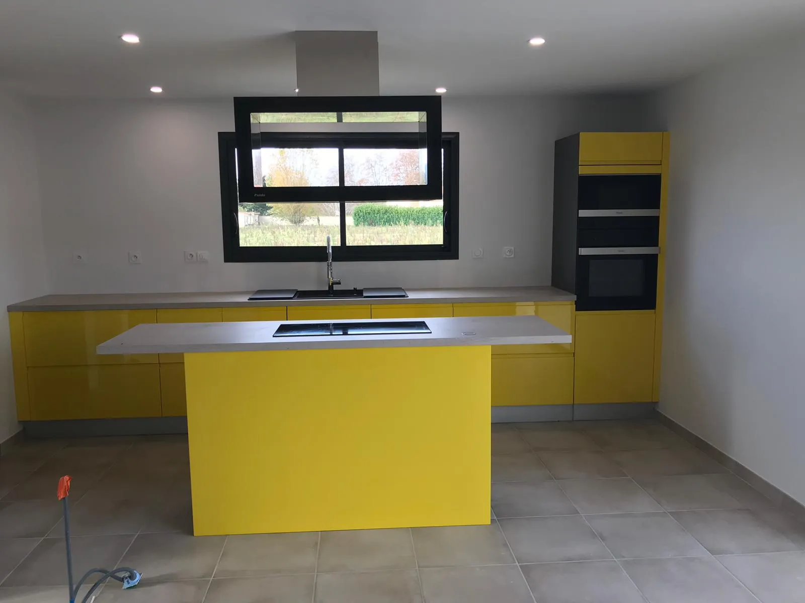 Cuisine façades jaune vif laquées, plan de travail béton, fenêtre noire ouverte sur la campagne — vue d'ensemble après livraison